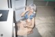 A woman with blue hair sitting on a bed next to a refrigerator.